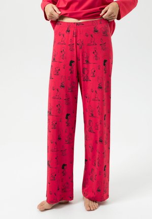 Rode pyjamabroek met zwart Peanuts-stripfigurenpatroon, blootvoets gedragen door een persoon die een rode top optilt, tegen een effen witte achtergrond.