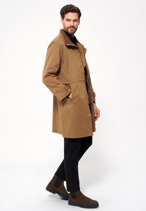 Brązowy wodoodporny płaszcz z wysokim kołnierzem, przednimi kieszeniami i zapięciem na guziki. Połączony z czarnymi spodniami i brązowymi botkami za kostkę.
