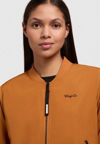 Oranje bomberjack met ronde kraag, zwarte rits en logodetails. Gladde textuur en aansluitend ontwerp met contrasterende zwarte accenten.