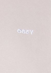 Ljust rosa tyg med en broderad "OBEY"-logotyp i vitt. Slät textur med ett ribbat ytmönster.