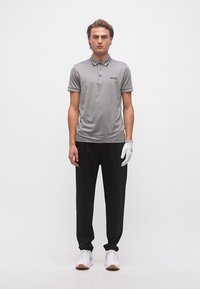 Νεαρός άνδρας που φορά γκρι πόλο μπλουζάκι με το λογότυπο "BOSS", μαύρο παντελόνι, άσπρα αθλητικά παπούτσια και λευκό γάντι στο δεξί του χέρι, στέκεται μπροστά σε λευκό φόντο.