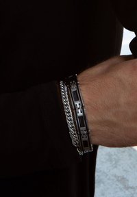 Zwarte en zilveren armbanden gestapeld op een pols; de ene is een geometrisch metalen ontwerp met een patroon, de andere is een chunky zilveren ketting.
