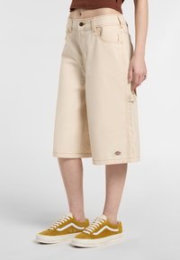 Pantaloni cargo beige in cotone, con taglio dritto, tasche laterali e cuciture arancioni, abbinati a scarpe da ginnastica gialle e pelose.
