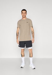 Beige kortermet skjorte med en subtil tekstur og svart logo, kombinert med mørke shorts og hvite sneakers. Hud synlig med tatoveringer.