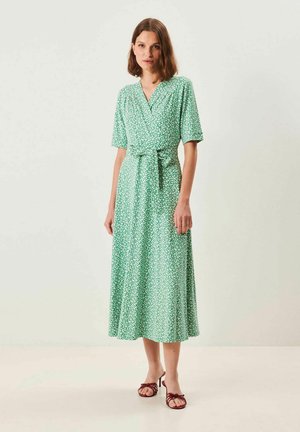 Nainen seisoo käyttäen vihreää, kukallista kietaisumekkoa, jossa on lyhyet hihat ja joka on solmittu vyötäröltä, sekä ruskeita avonaisia korkokenkiä.