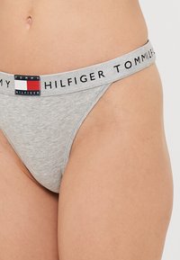 Sivý bavlnený tangá s čiernym pásom, na ktorom je logo "Tommy Hilfiger" a malá obdĺžniková visačka značky v červenej, bielej a modrej farbe.