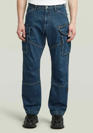 Pantaloni cargo in denim con più tasche grandi e dettagli con cerniere, indossati con sneakers nere e una maglietta bianca.