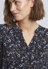 Blusa floreale in blu navy con accenti bianchi e dorati. Caratterizzata da un scollo a V e un tessuto leggero e testurizzato con un motivo di piccoli fiori.