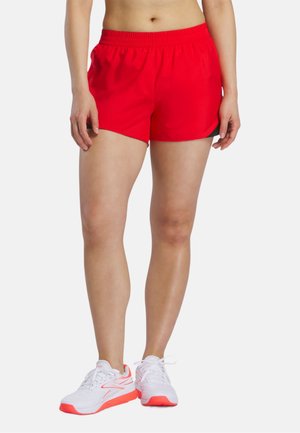 Frau trägt leuchtend rote Sportshorts und weiße Laufschuhe mit roten Akzenten, steht vor einem schlichten hellen Hintergrund.