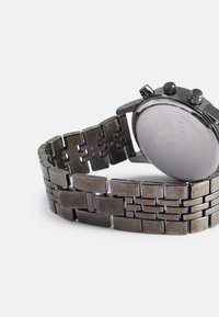 Edelstahl-Chronograph mit einem dunklen, metallischen Gliederarmband und einem glatten, runden silbernen Zifferblatt. Detaillierte Hardware-Elemente sind sichtbar.