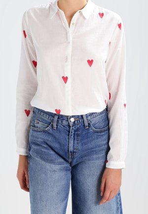 Chemise blanche boutonnée avec des imprimés de cœurs rouges, fabriquée en tissu léger. Associée à un jean denim bleu taille haute, avec une coupe classique.