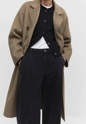 Personne portant un long manteau beige, un cardigan noir boutonné sur une chemise blanche et un pantalon noir taille haute avec les mains dans les poches.