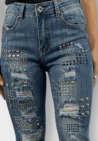 Jeans de mezclilla azul de cintura alta que presentan secciones desgastadas, tachuelas plateadas y patrones geométricos para añadir textura y detalle.