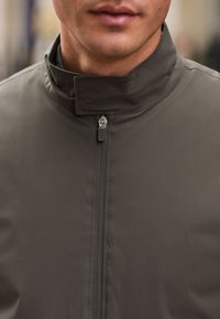 Graue Jacke mit Reißverschluss und hohem Kragen, die eine glatte Textur und ein minimalistisches Design aufweist. Die Reißverschlussbeschläge sind deutlich sichtbar.