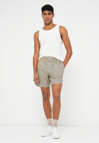 Uomo in piedi con una canottiera bianca, pantaloni bermuda beige a fantasia, calze beige e sneakers bianche su uno sfondo bianco semplice.