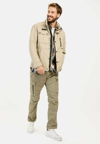 Jasno beżowa kurtka z wieloma kieszeniami, noszona na kraciastą koszulę. Oliwkowe spodnie z kieszeniami typu cargo. Białe trampki dopełniają stylizację.