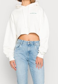 Witte cropped hoodie met capuchon, zachte stof, losse pasvorm en groene tekst "COLOURFUL REBEL". Gecombineerd met lichtblauwe, hooggetailleerde spijkerbroek.