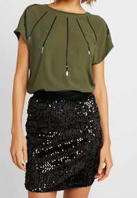 Blusa verde oliva de manga corta con acentos de lentejuelas negras y metálicas; combinada con una falda corta negra de lentejuelas con textura fruncida.