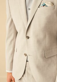 Veste de costume grise clair sur mesure avec deux boutons, poche avant contenant un mouchoir à motifs, assortie à une chemise légèrement à motifs.