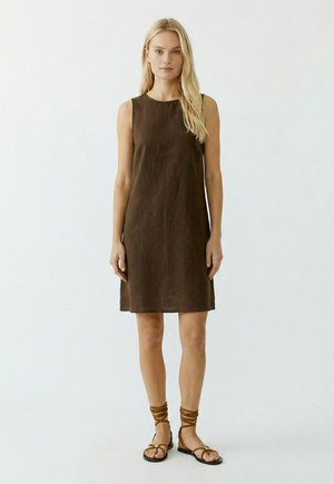 Frau mit langen blonden Haaren, die ein ärmelloses, braunes Kleid knielanger Länge und braune, flache Sandalen mit Riemchen trägt, steht vor einem schlichten, hellen Hintergrund.