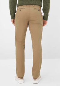 Beige Baumwollhosen mit geradem Schnitt, ausgestattet mit Gesäßtaschen mit Knopfverschluss. Kombiniert mit weißen Sneakers und einem grünen Pullover.