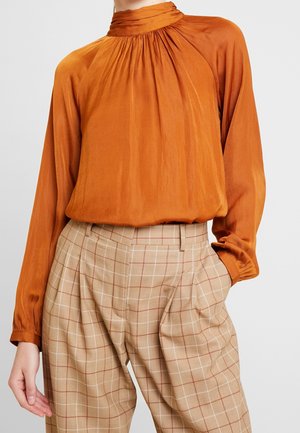 Rustrød bluse med høj hals og lange ærmer kombineret med beige ternede plisserede bukser, båret af en stående person med den ene hånd i lommen.