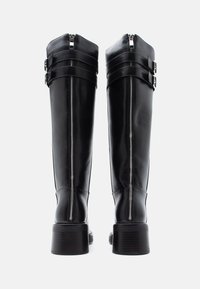 Μαύρες δερμάτινες μπότες μέχρι το γόνατο με γυαλιστερό φινίρισμα, με φερμουάρ στο πίσω μέρος, δύο ρυθμιζόμενα λουριά και χοντρούς τακούνια.