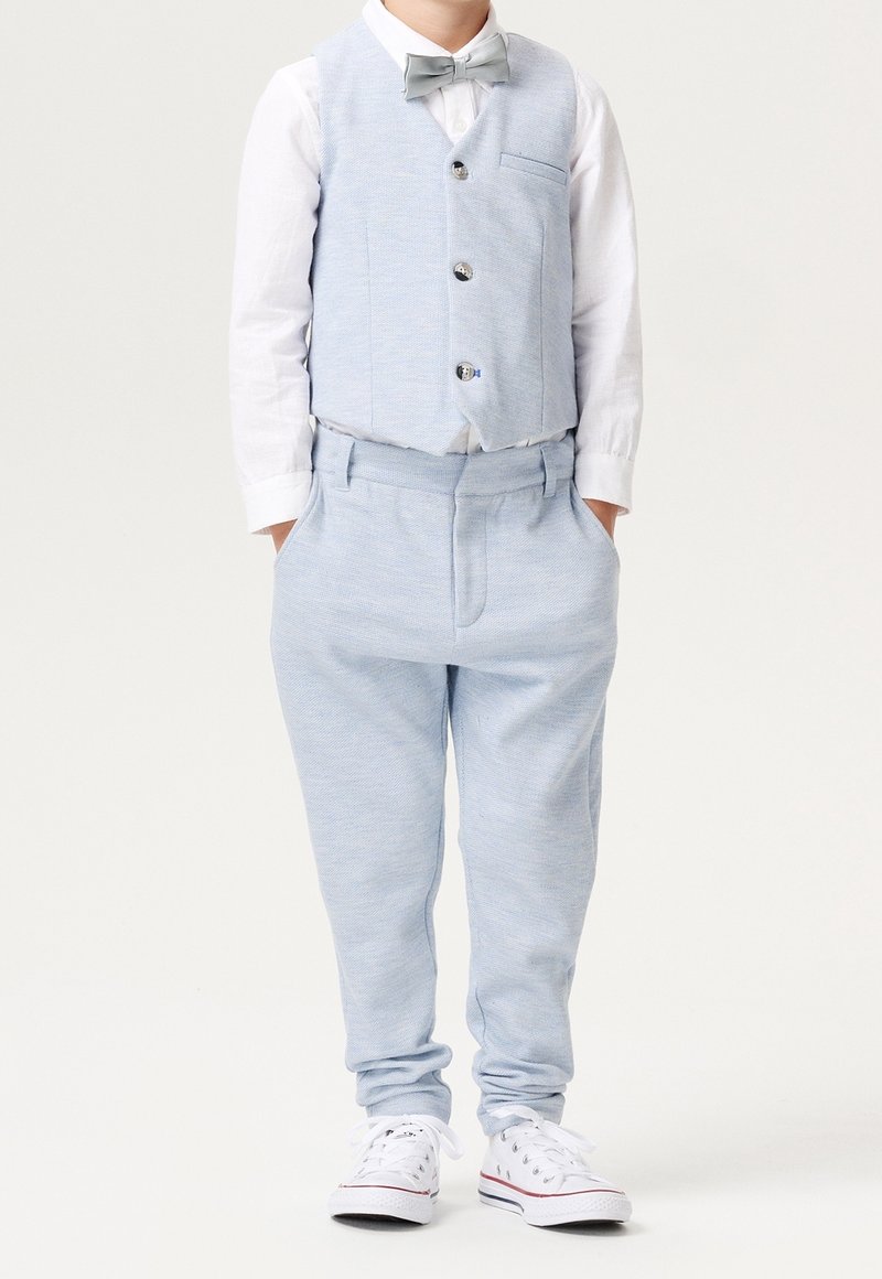 Jeune garçon portant un gilet bleu clair, un pantalon assorti, une chemise blanche, un nœud papillon gris et des baskets blanches, les mains dans les poches.