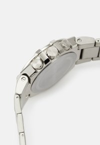 Edelstahl-Uhr mit Metallarmband, ausgestattet mit Drückern, einer strukturierten Lünette und einem glatten Gehäuseboden. Silberfarbe mit polierten Akzenten.