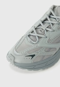 Tenis gri deschis, fabricat din plasă și material sintetic, cu o talpă texturată, modele curbe distincte și șireturi tonale.