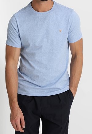 Hombre vestido con una camiseta de cuello redondo de manga corta azul clara con un pequeño emblema dorado en el pecho y pantalones negros, de pie con una mano en el bolsillo.