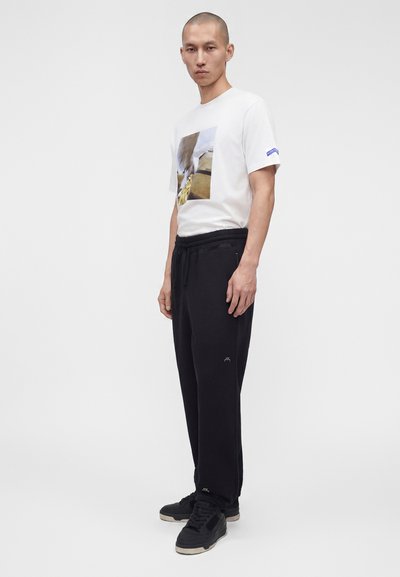 Νέος άντρας που φοράει λευκό γραφικό t-shirt, μαύρο παντελόνι με κορδόνι και μαύρα αθλητικά παπούτσια, στέκεται μπροστά σε ένα απλό φωτεινό φόντο.