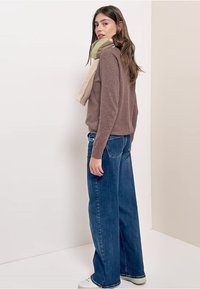 Brauner Pullover, weit geschnittene blaue Jeans und ein buntes Halstuch, das über die Schulter gelegt ist. Weiße Turnschuhe. Einfacher, lässiger Stil.