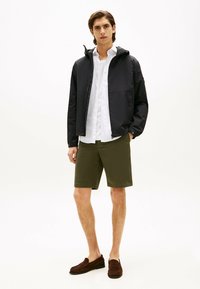 Chaqueta negra con capucha sobre una camisa blanca de botones, combinada con pantalones cortos verde oliva y mocasines marrones. Diseño minimalista y líneas limpias.