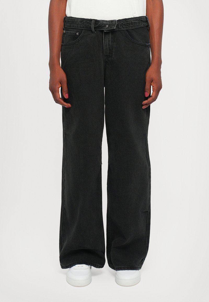 Jeans anchos negros con un acabado texturizado, que presentan un cierre de botón y cremallera, presillas para el cinturón y bolsillos delanteros. Zapatillas blancas en la parte inferior.