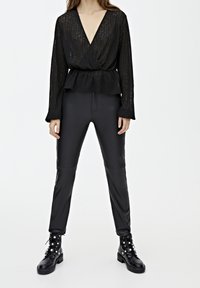 Blouse noire transparente à manches longues et décolleté en V profond, associée à un pantalon noir en simili cuir ajusté et des bottes de combat à clous argentés.