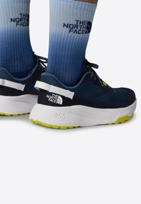 Modré sportovní boty s texturovaným svrškem, bílou mezipodešví a žlutou podrážkou. Nahoře nad botami jsou vidět šedé a modré gradientové ponožky.