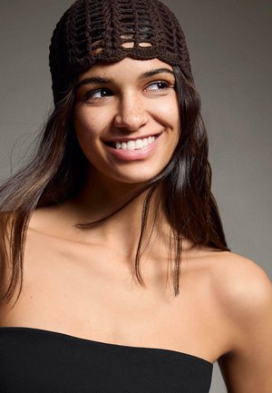 Jonge vrouw met lang donker haar, die een bruine gehaakte muts en een zwarte strapless top draagt, glimlacht en kijkt naar de zijkant.