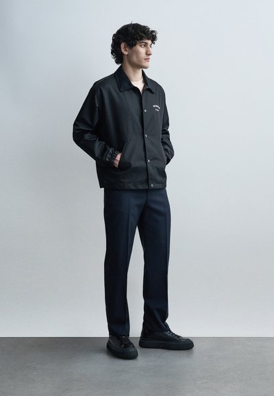 Νέος άνδρας όρθιος με τα χέρια στις τσέπες, φορώντας μαύρο σακάκι με κουμπιά, σκούρο μπλε παντελόνι και μαύρα αθλητικά παπούτσια, μπροστά σε απλό φωτεινό φόντο.