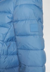 Rękaw jasnoniebieskiej kurtki puchowej o pikowanej fakturze, z kwadratową gumową naszywką z napisem "BOSS" i kontrastującymi detalami przeszyć.