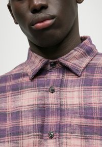 Fioletowa i różowa koszula flanelowa w kratę z kołnierzykiem zapinanym na guziki i czarnymi guzikami, o miękkiej strukturze i symetrycznym wzorze.