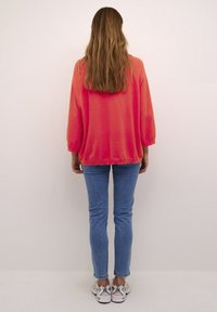 Koralfarbener übergroßer Sweatshirt mit Dreiviertelärmeln, kombiniert mit blauen Skinny-Jeans und weißen Sneakern mit silbernen Akzenten. Die Haare fallen bis zur Mitte des Rückens.