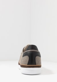 Sneaker in tessuto grigio con accenti in pelle nera, suola bianca e dettagli texturizzati marroni lungo il bordo inferiore. Design semplice e moderno.