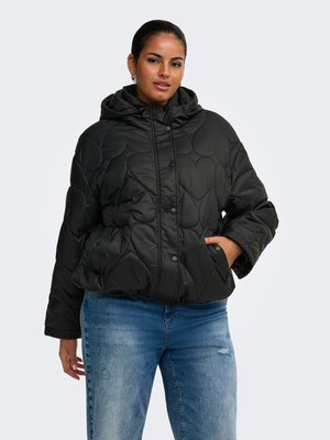 Frau trägt eine schwarze gesteppte Jacke mit Herzmustern und blauen Jeans, steht mit einer Hand in der Jackentasche vor einem schlichten Hintergrund.