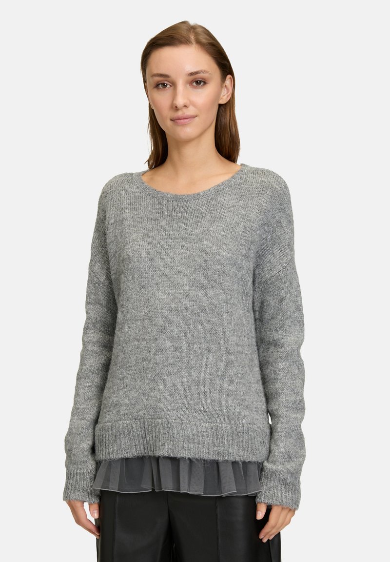 Un pull gris au fit ample, avec un col côtelé et des manches longues, superposé à un haut gris transparent et à volants. Texture en tricot douce.