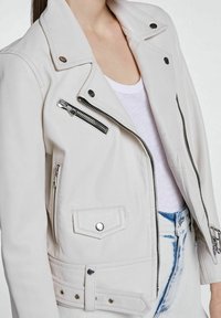 Jasny beżowy kurtka motocyklowa z srebrnymi zamkami błyskawicznymi, zatrzaskami i usztywnianym kołnierzem. Posiada przednie kieszenie i fakturowaną tkaninę.