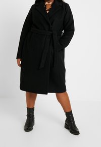 Abrigo negro de lana con cinturón, cuello con muesca, mangas largas y bolsillos laterales. Llevado sobre una blusa estampada y combinado con botas negras por el tobillo.