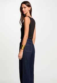 Top noir sans manches avec une finition texturée, associé à un pantalon en denim foncé taille haute. Un bracelet en métal doré accentue le look.