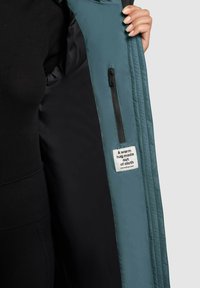 Zielonkawe zewnętrzne kurtka pikowana, wyposażona w czarną podszewkę, kieszeń na zamek i materiałową metkę z napisem „Ciepły uścisk wykonany z tkaniny.”