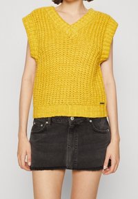 Geel gebreid mouwloos topje met een V-hals, voorzien van een textuurpatroon en een geribbelde zoom, gecombineerd met een zwarte denim mini rok.